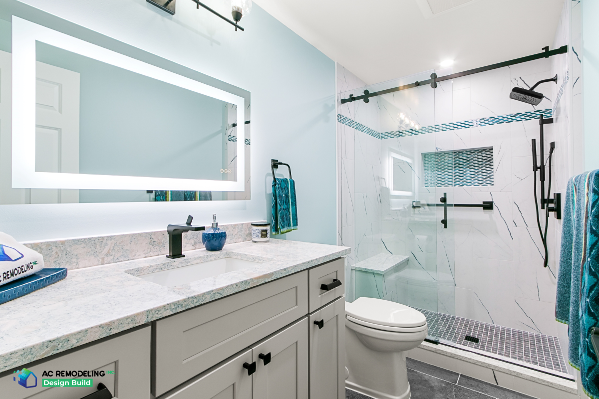 Remodeled Gaithersburg bathroom with backlit mirror, gray shaker vanity, quartz top and marble-look shower with blue glass accent tile